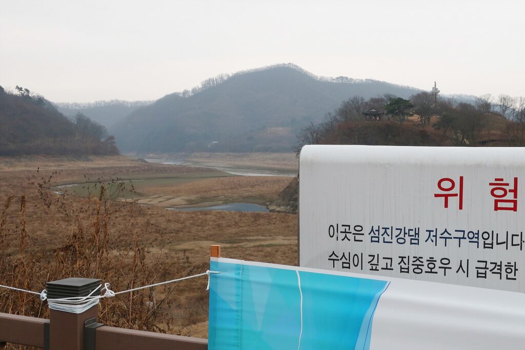 [오른쪽 아래에 경고 표지판] 위험: 이곳은 섬진강댐 저수구역입니다 수심이 깊고 집중호우 시 급격한(이하 가려짐) 경고 표지판 뒤로 물이 완전히 빠진 옥정호 기슭이 보인다.