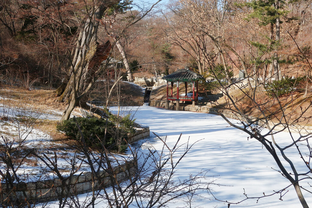 하얀 잔설이 쌓인 연못과 큰 가지가 꺾인 나무. 그리고 멀리 관람정이 보인다.