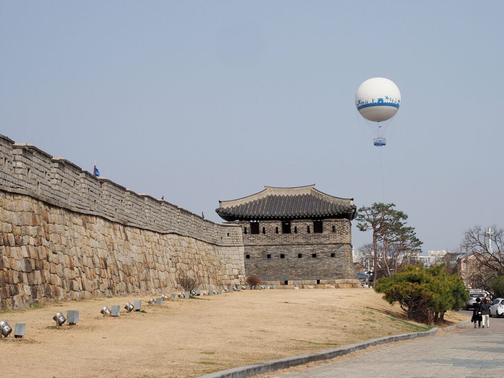 수원 화성 성벽이 곧게 뻗어 있고 동포루가 그 끝에 서 있다. 뒤로는 하얀 열기구가 떠 있다.