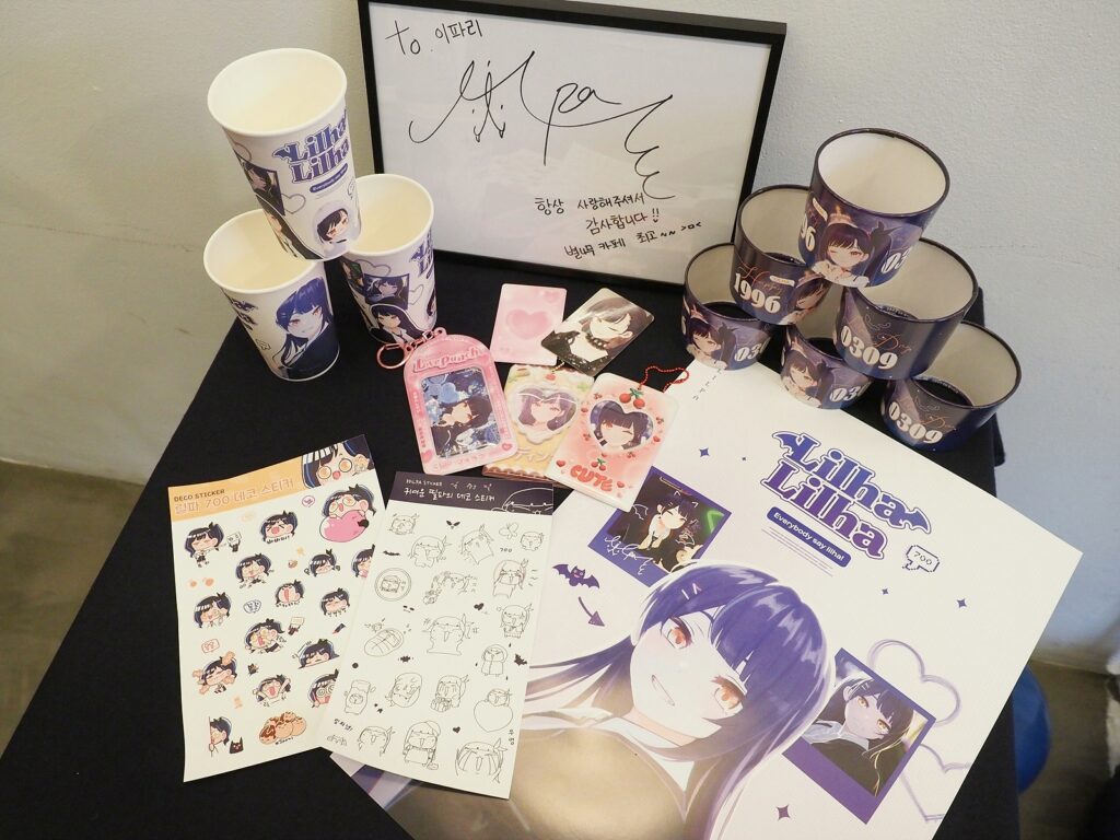 릴파 굿즈들. 컵홀더, 포스터, 스티커 등이 놓여 있고 뒤로는 릴파의 싸인이 액자에 들어 있다.