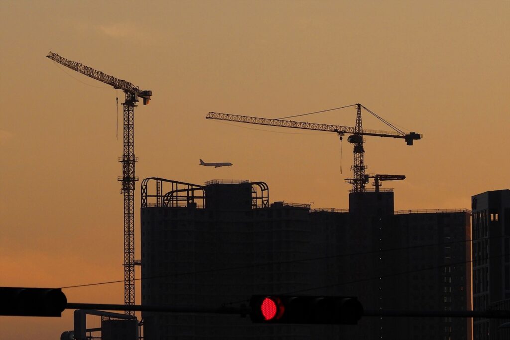 노랗게 물드는 노을을 배경으로 건설 중인 고층건물이 실루엣으로 비친다. 타워크레인 두 대 사이로 인천공항에 착륙하려 하는 비행기가 보인다.