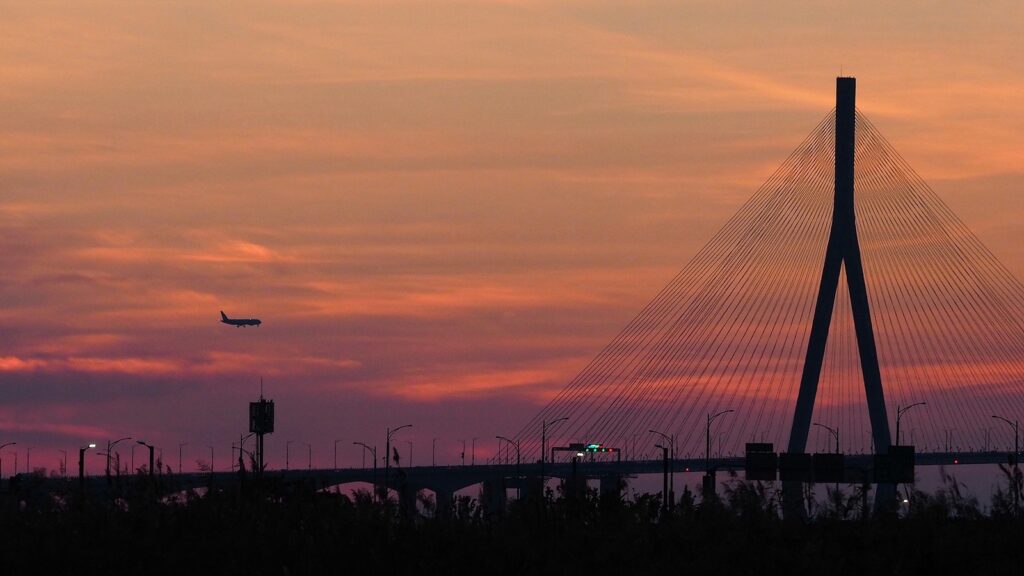 붉게 물드는 하늘 앞에 인천대교가 실루엣으로 보인다. 그 위로 비행기가 지나고 있다.