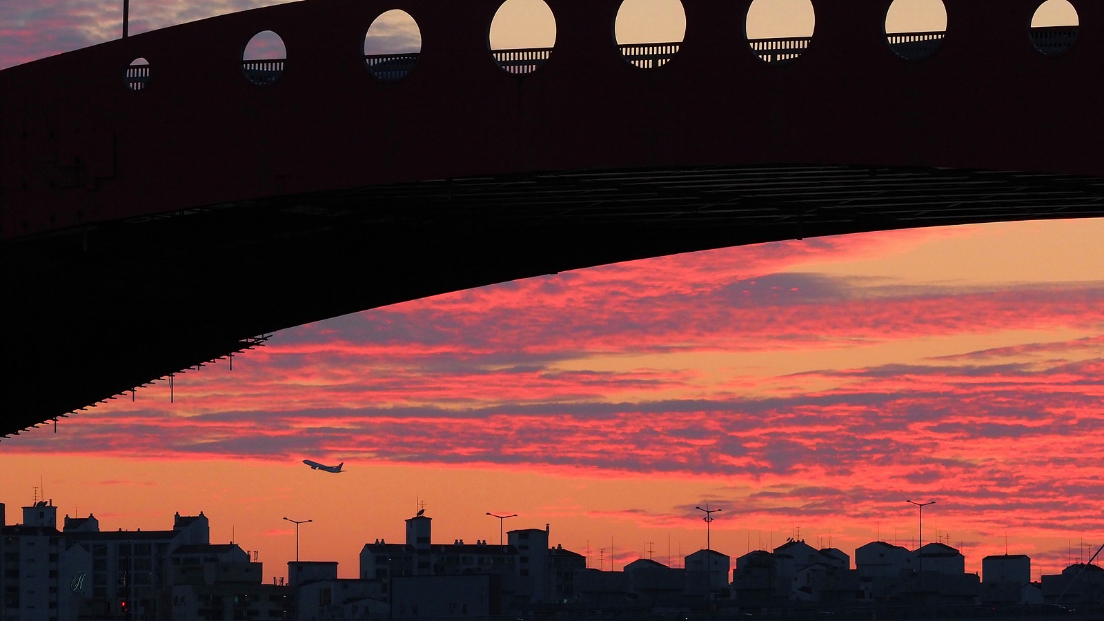 붉게 타는 하늘 아래로 한강 남쪽 건물들이, 위로 성산대교 구조물이 걸쳐 있다. 비행기 한 대가 건물 위로 고도를 높이며 날아가고 있다.