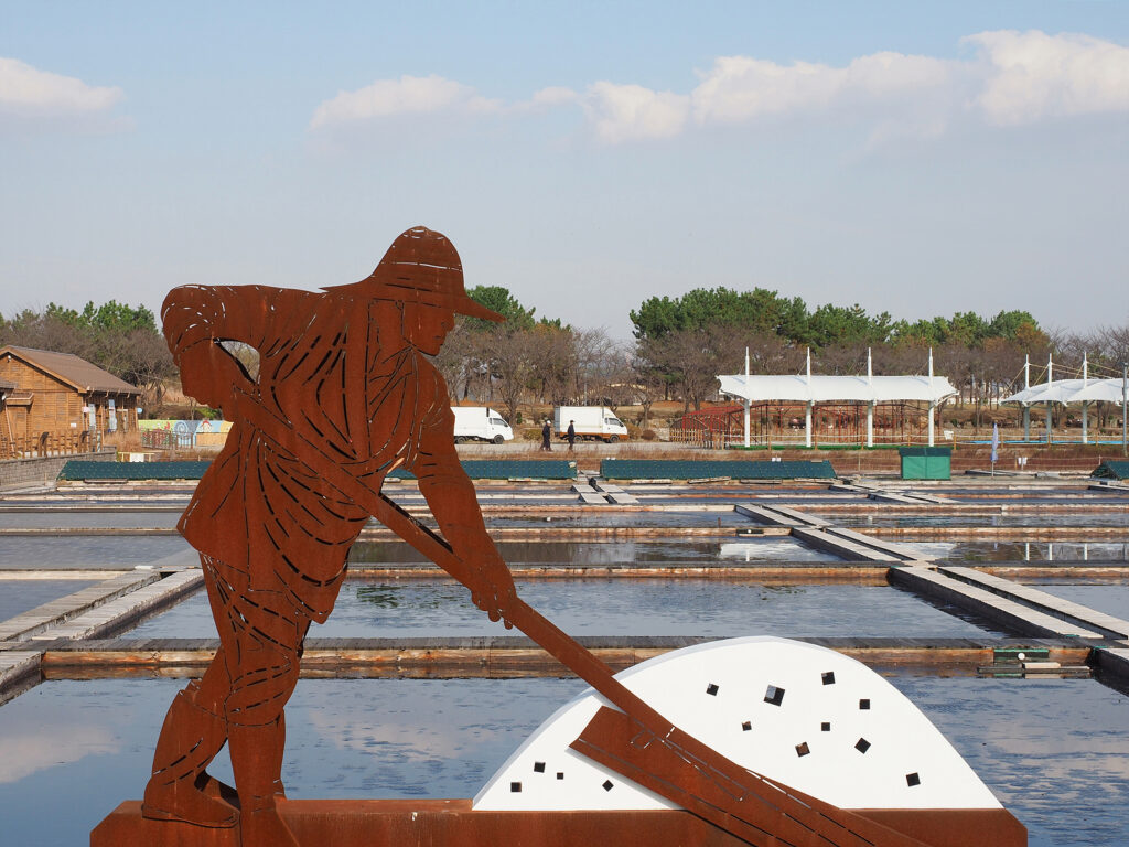고무래로 소금을 모으는 염부 모양의 붉은 금속제 조형물. 그 뒤로 네모난 염전이 보인다.
