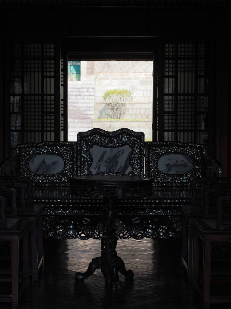 어두운 방 한가운데 놓인 자개 의자와 테이블. 그 뒤로 열린 문을 통해 밝은 언덕이 보인다.