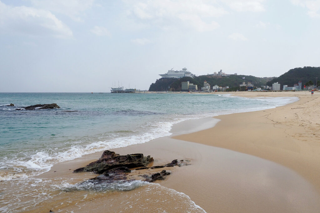 백사장이 펼쳐져 있는 가운데 푸른 바닷물이 밀려오고 있다. 정면 멀리, 커다란 배 모양의 리조트가 보인다.