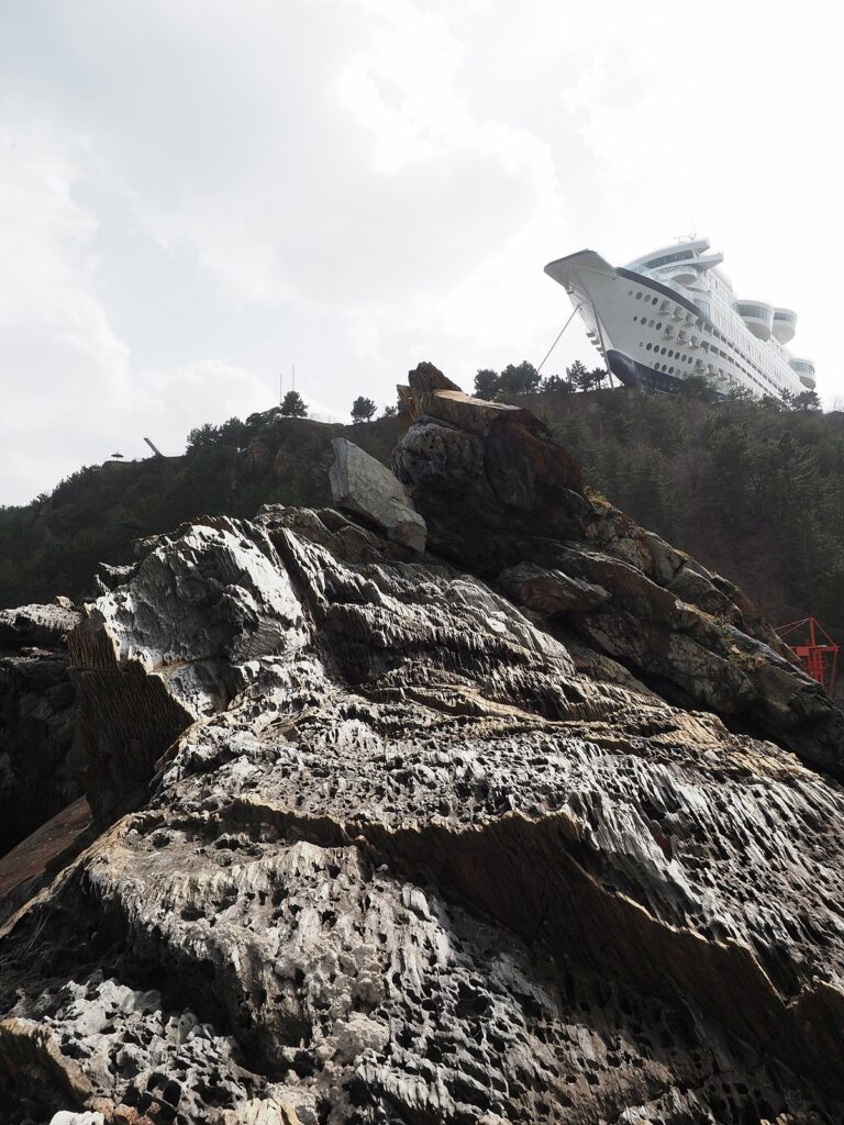 서 있는 바위, 그 뒤 산비탈을 지나 꼭대기에 커다란 배 모양의 건물이 서 있는 것을 로우 앵글로 바라본 모습.