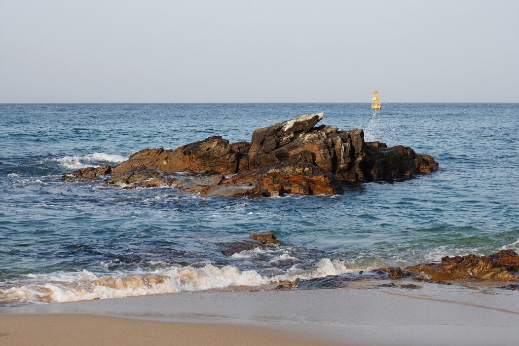 파도가 잔잔하게 밀려오는 해변. 몇 걸음 앞에는 바위섬이 하나 떠 있다. 전체적으로 늦은 오후의 오렌지빛 햇살이 섞여 있다.