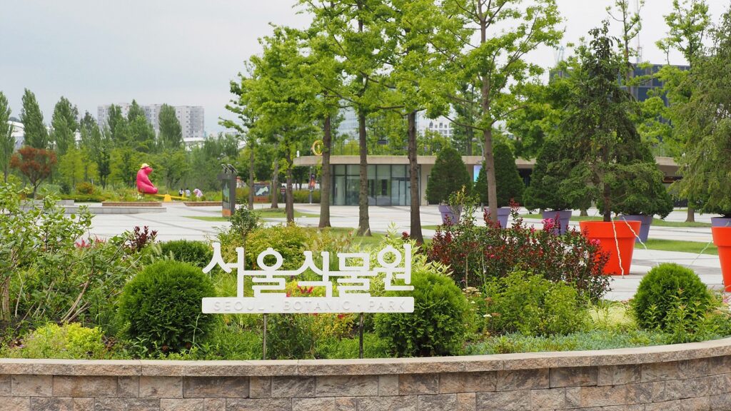 '서울식물원'이라 적힌 팻말이 서 있고 뒤로 나무들이 늘어서 있다. 곰 모양의 조형물도 하나 보인다.