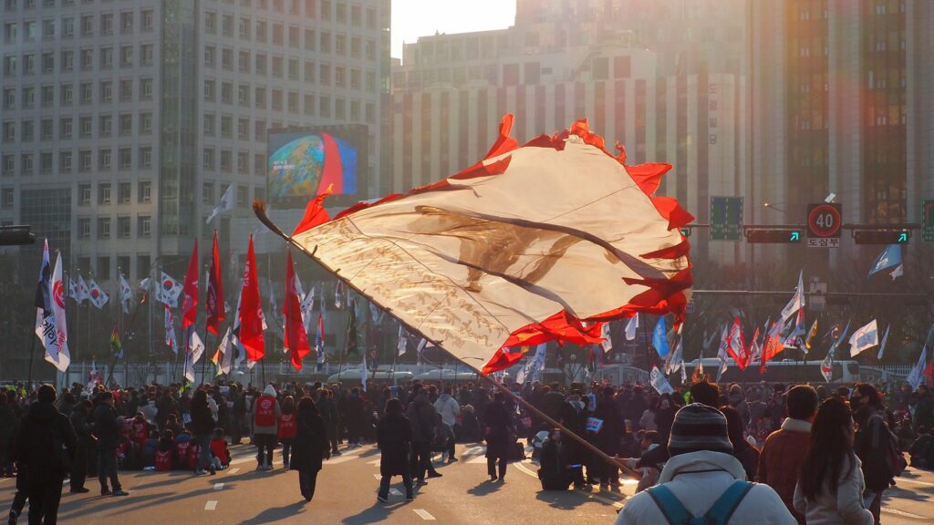 시민들이 모여 있는 가운데 그 사이에서 초대형 깃발이 나부끼는 해질녘의 광장.