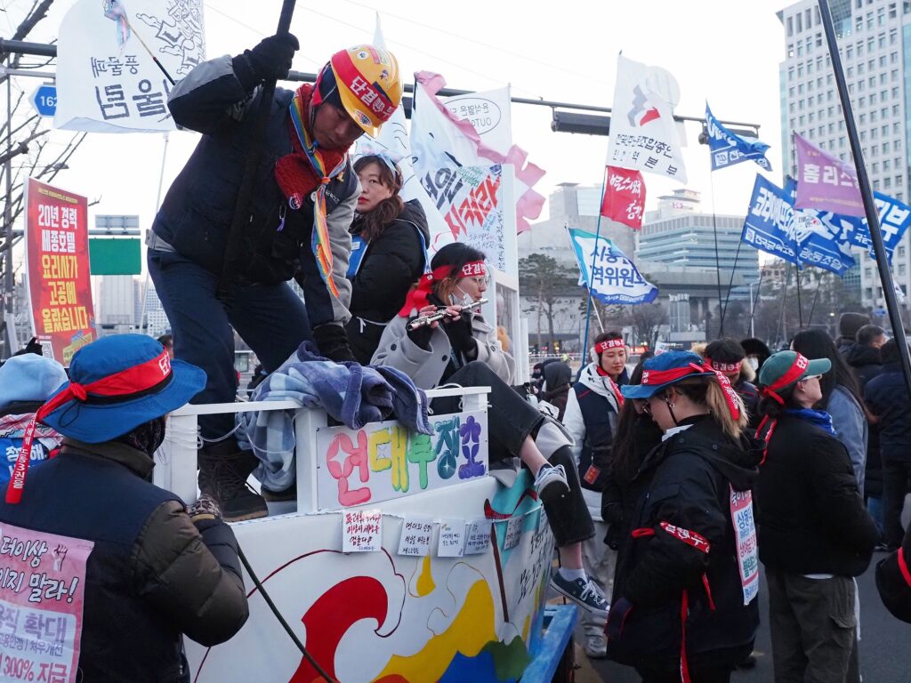 "연대투쟁호"라고 적힌 배 모양의 구조물 주변에 시민들이 모여 있다. 한 시민은 배에 걸터앉아 플루트를 불고 있다.