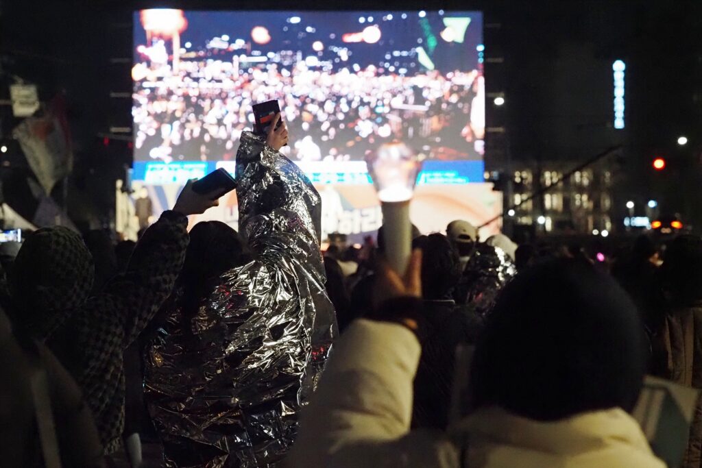 은박 담요를 두른 시민을 포함해 여러 시민이 응원봉을 들고 소리치는 뒷모습.