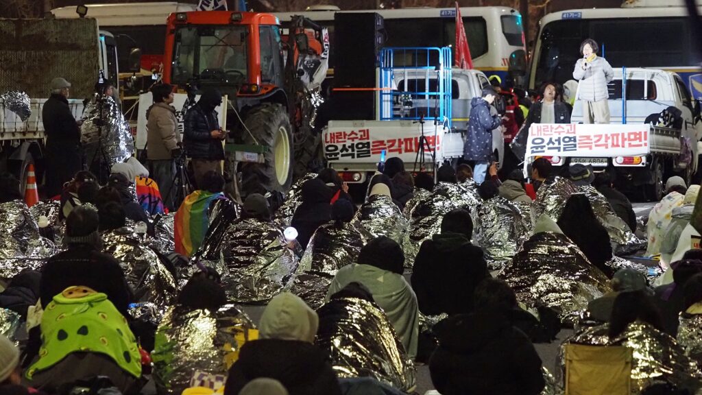 은박 담요를 뒤집어 쓴 시민들이 앉아 있고, 트랙터와 1톤 트럭이 맨 앞에 있다. 트럭 위에서 한 시민이 발언하고 있다.