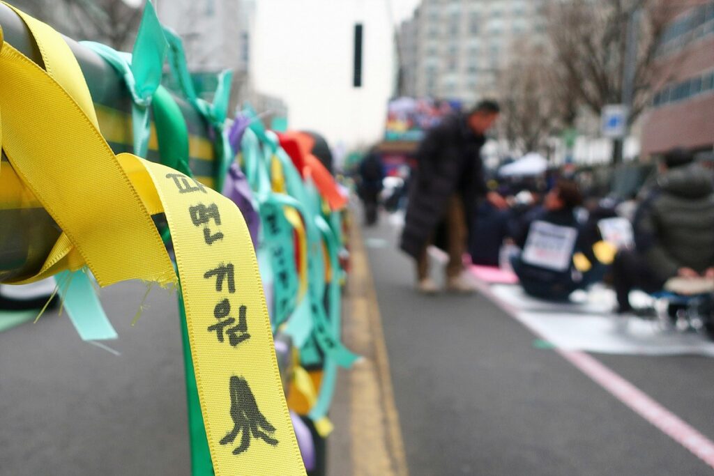 안국역 앞 도로. 중앙분리대에 "파면 기원" 등의 문구가 적힌 노란 리본, 녹색 리본, 무지개 리본 등이 묶여 있다. 도로에는 시민들이 연좌 중이다.