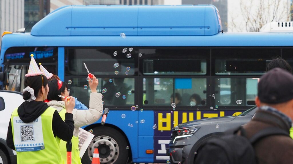 파란 시내버스가 지나는 앞으로 비상행동 스태프들이 고깔모자를 쓴 채 비눗방울을 불고 있다.