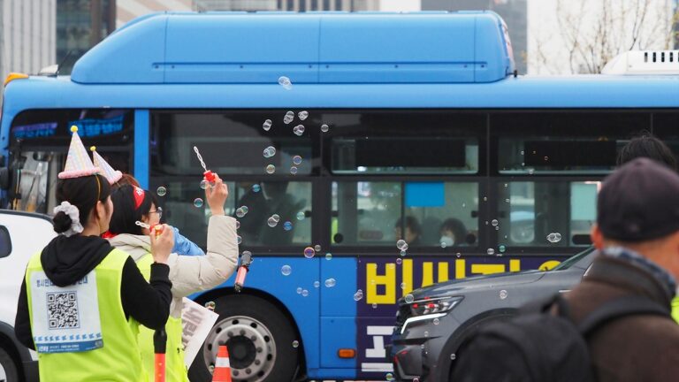 파란 시내버스가 지나는 앞으로 비상행동 스태프들이 고깔모자를 쓴 채 비눗방울을 불고 있다.
