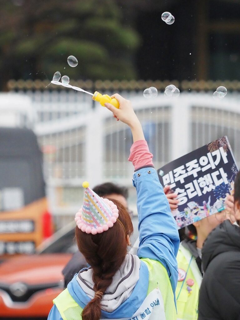 "민주주의가 승리했다"는 문구가 적힌 손팻말을 든 시민 앞에서 한 비상행동 측 스태프가 고깔모자를 쓴 채 비눗방울을 만들고 있다.