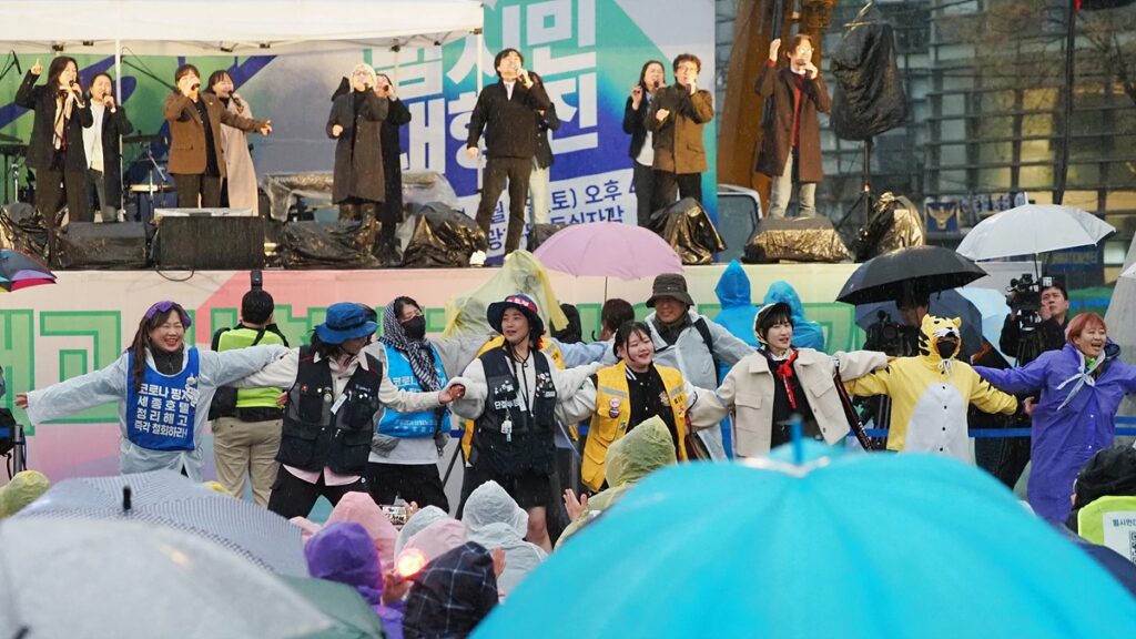 비가 내려 우산과 우비를 갖춘 시민들이 모여 있는 가운데, 무대 위와 무대 아래에 비상행동 측 공연자, 스태프 등이 노래와 춤으로 승리의 기쁨을 나누고 있다.