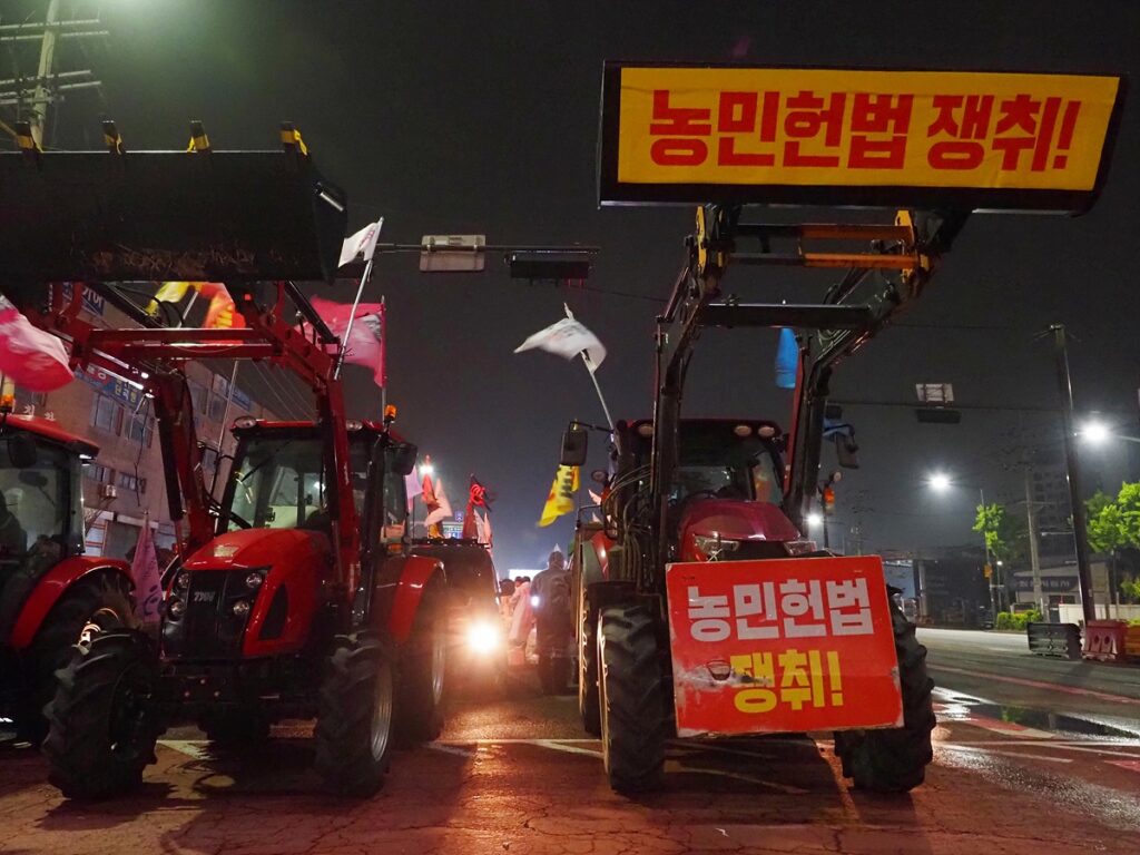 "농민헌법 쟁취!"라는 문구가 적힌 팻말을 붙인 트랙터가 멈춰 서 있다. 그 뒤로 시민들의 깃발이 나부낀다.
