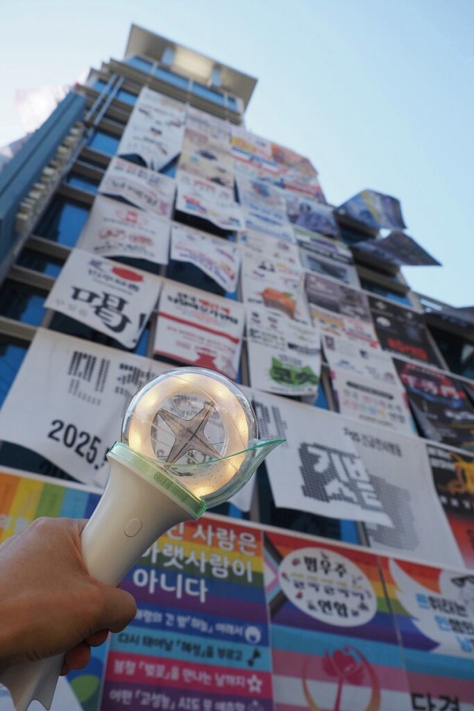 건물 외벽에 수많은 깃발이 걸려 있다. 왼쪽 아래엔 노란 빛을 내는 응원봉을 든 손이 보인다.